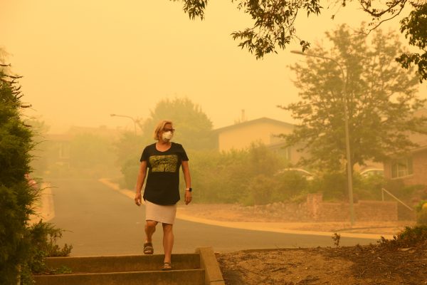 A woman walks in the street and wears a face protective mask. Everything is very dry and there is a dangerous level of air pollution. A thick yellow fog coming from the nearby forest fires covers Canberra city. Wildfire particulate pollution